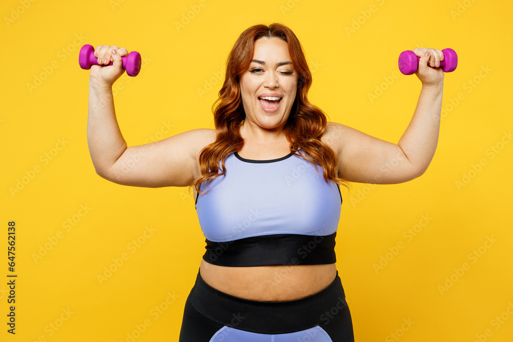 Young fun happy chubby overweight plus size big fat fit woman wear blue top warm up training hold dumbbells wink look camera isolated on plain yellow background studio home gym. Workout sport concept. Young fun happy chubby overweight plus size big fat fit woman wear blue top warm up training hold dumbbells wink look camera isolated on plain yellow background studio home gym. Workout sport concept.