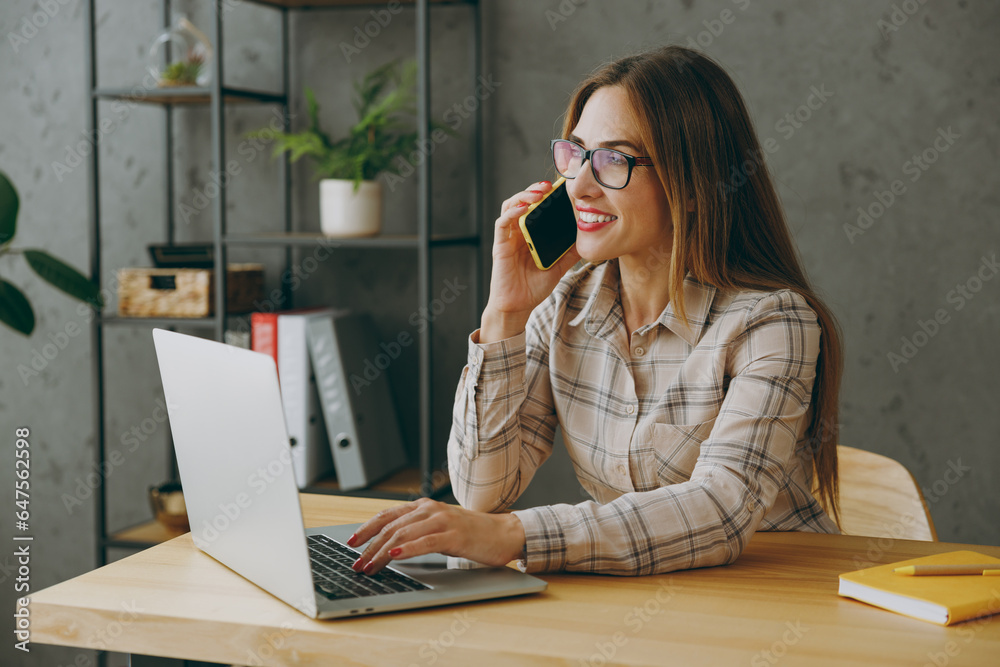 Side view young successful employee business woman wearing shirt casual clothes glasses sit work at office desk with pc laptop computer talk speak on mobile cell phone. Achievement career job concept. Side view young successful employee business woman wearing shirt casual clothes glasses sit work at office desk with pc laptop computer talk speak on mobile cell phone. Achievement career job concept.