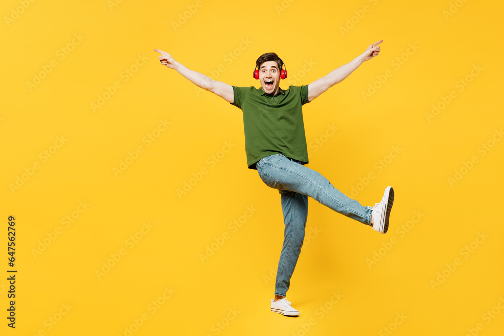 Full body side view young happy man he wears green t-shirt casual clothes listen to music in headphones raise up hands leg dance isolated on plain yellow background studio portrait. Lifestyle concept.