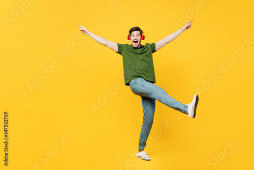 Full body side view young happy man he wears green t-shirt casual clothes listen to music in headphones raise up hands leg dance isolated on plain yellow background studio portrait. Lifestyle concept.