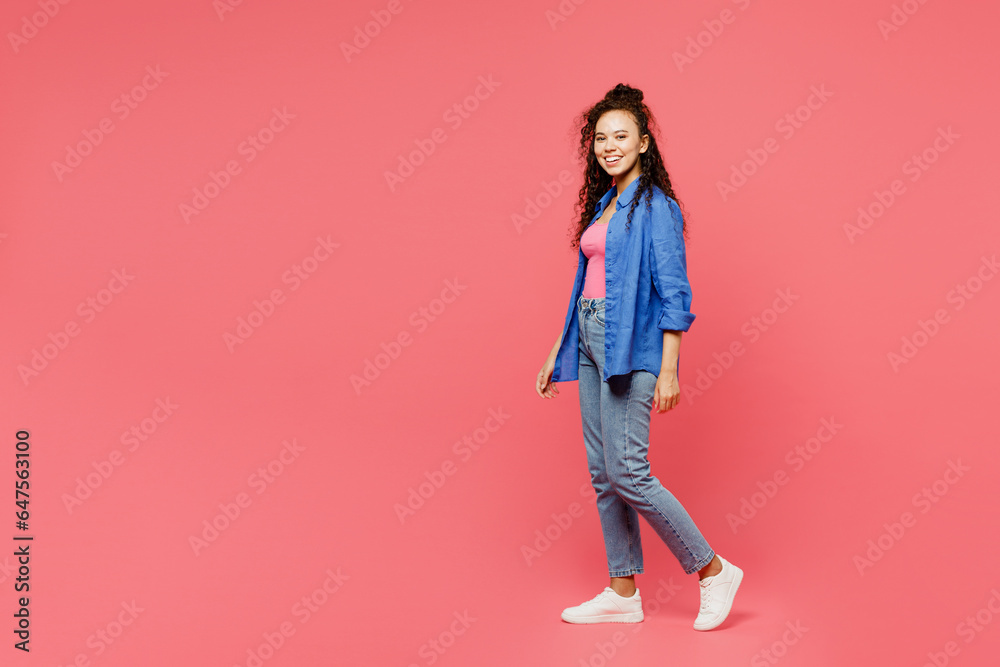 Full body side profile view happy young woman of African American ethnicity she wear blue shirt casual clothes walk go look camera isolated on plain pastel pink background studio. Lifestyle concept.