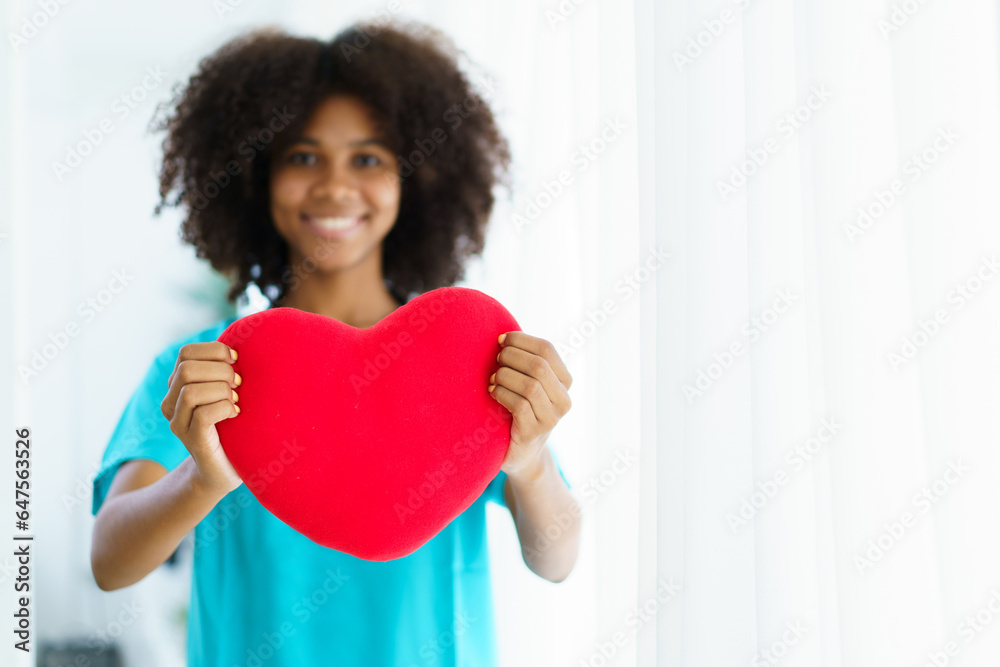 Happy cheerful African - American ethnicity female doctor holding and showing a red heart object to camera with copyspace. The Red Cross and giving concept. Happy cheerful African - American ethnicity female doctor holding and showing a red heart object to camera with copyspace. The Red Cross and giving concept.