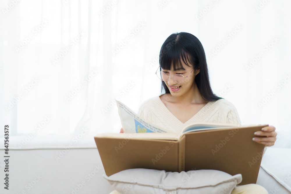 Happy - cheerful Asian young woman with eye glasses sitting on the sofa in living room and reading a book alone. Happy - cheerful Asian young woman with eye glasses sitting on the sofa in living room and reading a book alone.