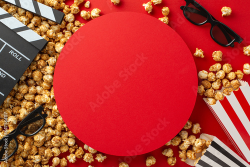 Silver Screen Magic: Overhead shot of popcorn, 3D glasses, and a clapperboard on red backdrop with empty circular advert space, the essential setup for a movie night with friends photo