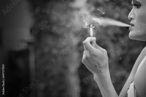 Black and white close up of Asian transwoman or transgender holding cigarette and smoking in garden. People, smoking and bad habits concept. Picture for World No Tobacco Day. Background or copy space. photo