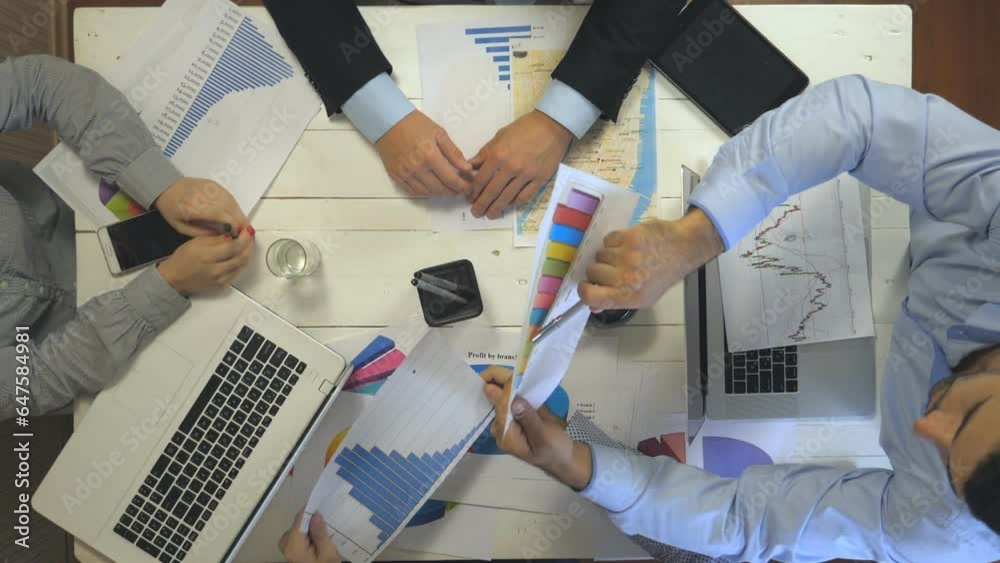 Top view to hands of business people sitting at table in trendy office and examining graphs. Group of busy employees working efficiently on business development. Creative team work indoor. Slow mo