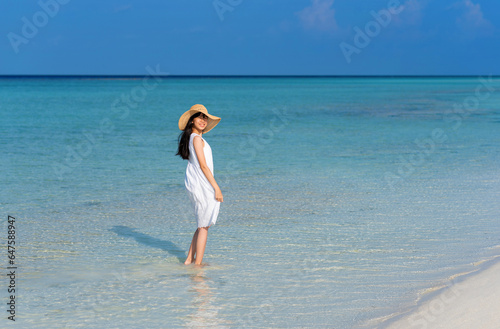 Smiling young Asian girl walking on white sand beach with clear blue sea and sky. Teenager girl wearing sun straw hat, white dress enjoy vacation. Outdoor summer travelling concept, copy space.