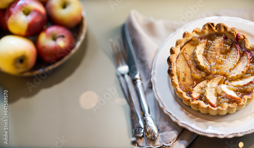 French Apple Tart baked with shortcrust pastry, also called Pâte Brisée, on table with cutlery and fresh apples, blurry bokeh lights for cozy Christmas holiday mood in background with copy space