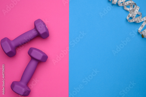 Layout of two rubberized dumbbells of 2kg purple color,measuring tape on a blue-pink background,top view.Sports training