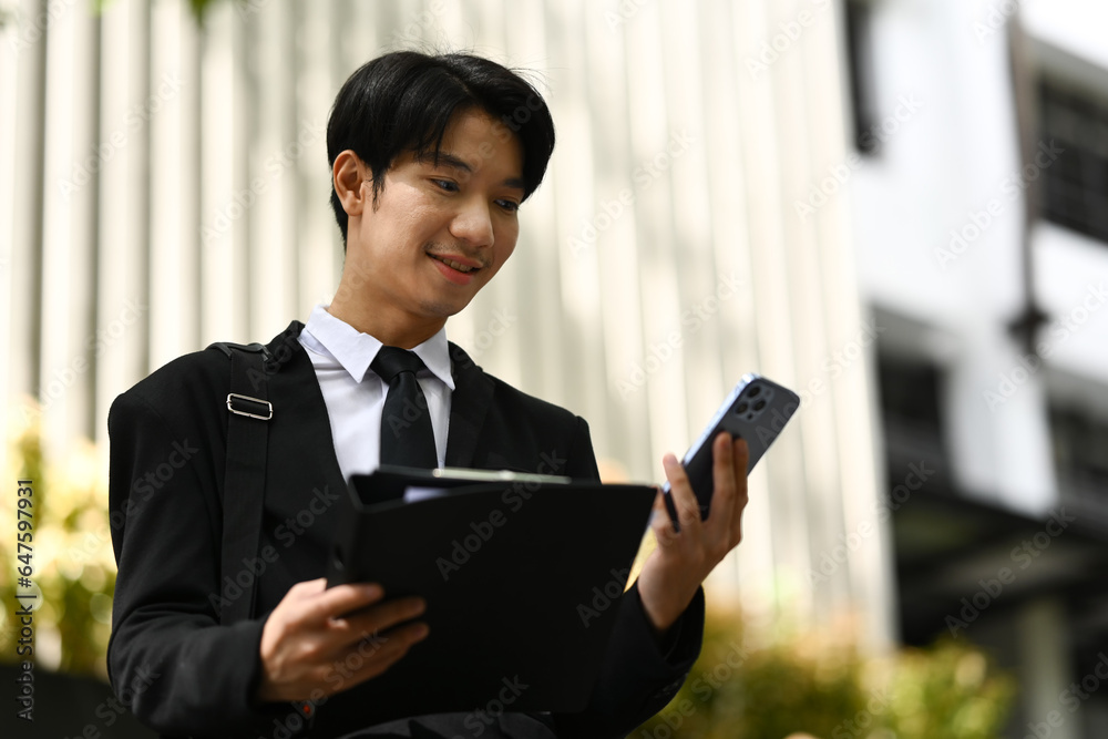 Young startup businessman using smartphone standing outdoors with office building in the background Young startup businessman using smartphone standing outdoors with office building in the background