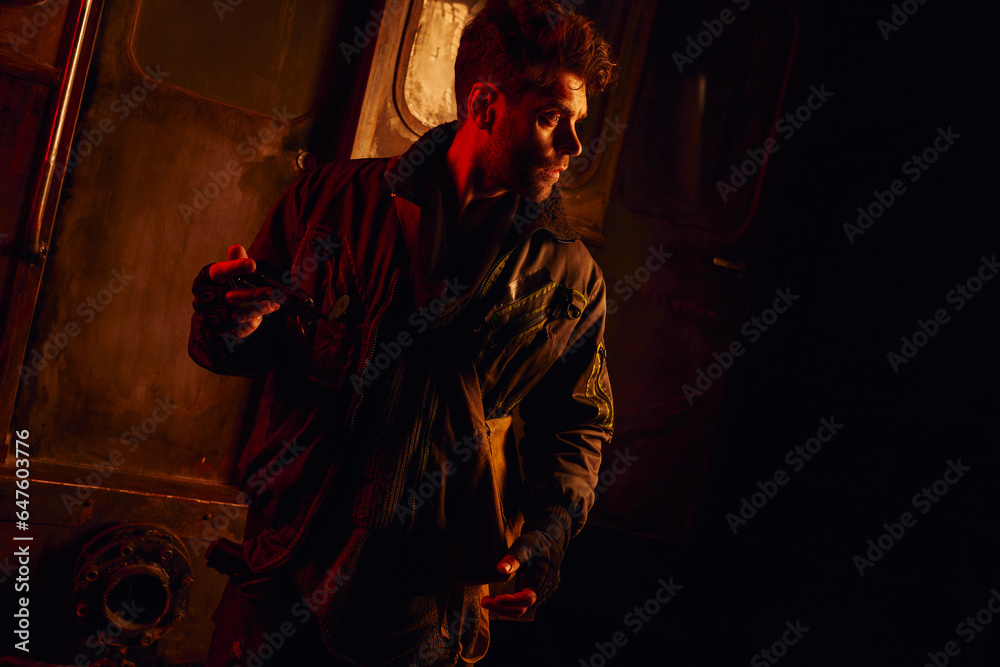 man holding spare part of subway carriage looking away in red light of post-disaster underground