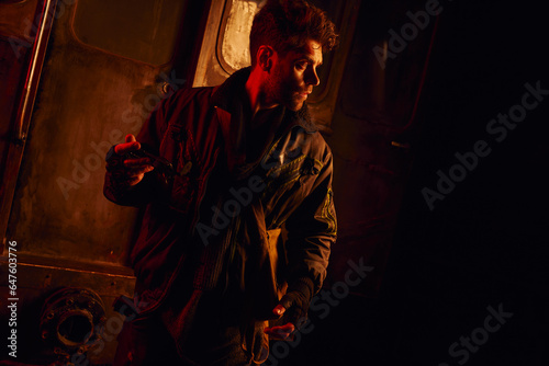 man holding spare part of subway carriage looking away in red light of post-disaster underground