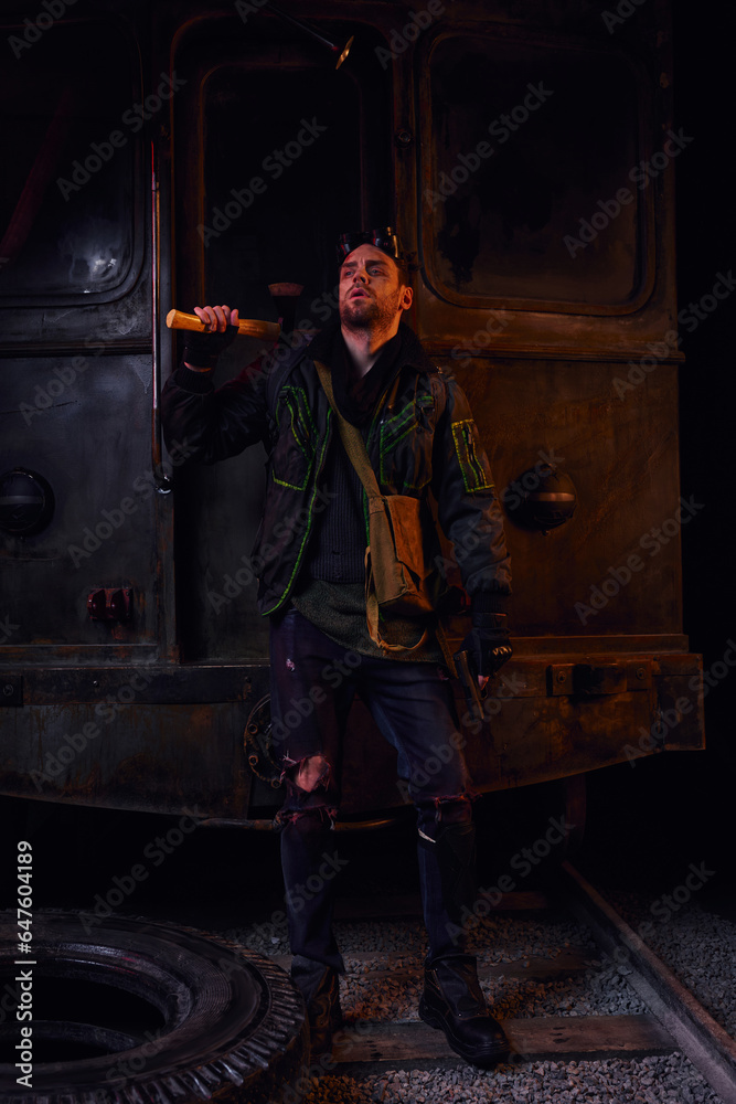man in worn dirty clothes standing with axe near rusty subway carriage, post-disaster survival