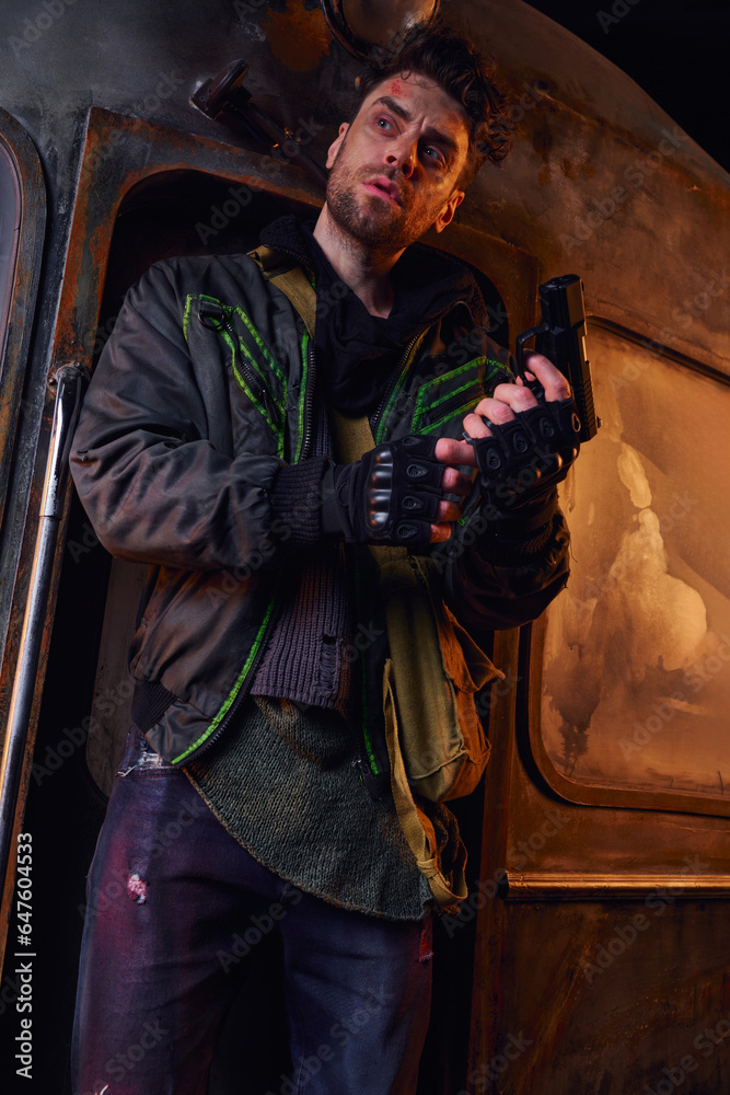 unshaven man in worn clothes and fingerless gloves holding gun and looking away in abandoned subway