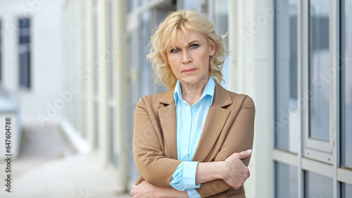 Tableau sur toile Strict serious mature blonde director crosses arms on chest standing against blurred company building
