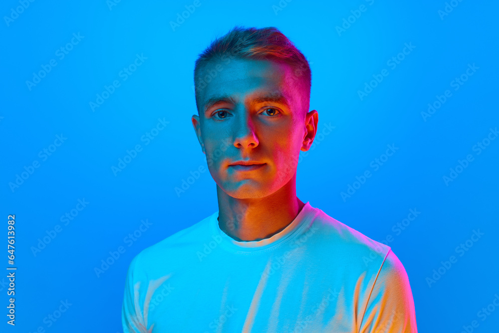 Portrait of young good-natured, calm man, student in white t-shirt looking at camera isolated on blue background in red neon light. Portrait of young good-natured, calm man, student in white t-shirt looking at camera isolated on blue background in red neon light.