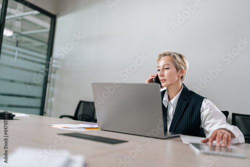 Remote low-angle view of gray-haired senior adult businesswoman talking on mobile phone at workplace table, sitting at laptop computer, looking to screen, discussing work, making call.