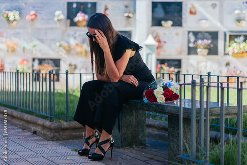 Canvas Print Weeping woman with sunglasses, black dress and hand on her head, sitting on a bench, with a bouquet of roses beside her, in a cemetery