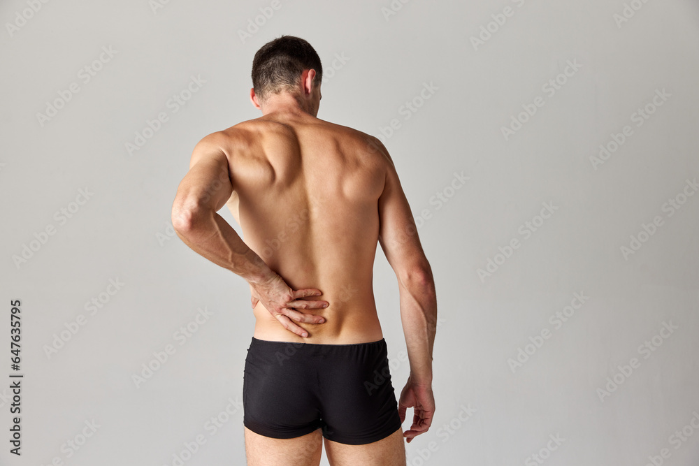 Back pains. Muscular man standing in underwear and holding his back against grey studio background. Medical treatment. Concept of men's health and beauty, body care, fitness, wellness, ad
