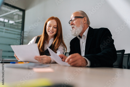 Low-angle view of bearded senior adult male CEO executive manager in formal wear mentoring smiling young female intern helping instructing trainee. Diverse-ages group discussing business plan together