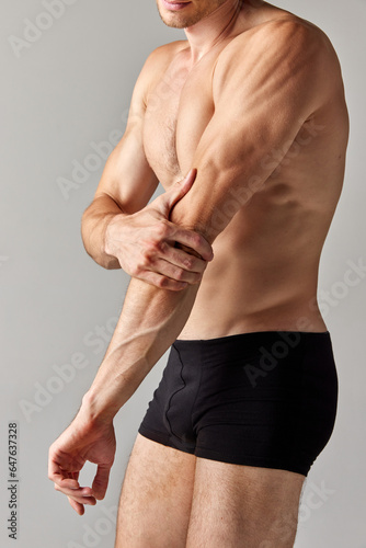 Hand pain  sprain. Cropped image of muscular man with relief body in underwear holding elbow against grey studio background. Concept of men s health and beauty  body care  fitness  wellness  ad