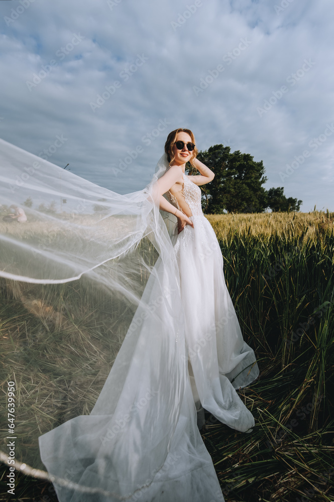 A beautiful young blonde bride in a white dress with a long veil in sunglasses stands in a wheat field outdoors in Ukraine. Wedding photography, portrait. A beautiful young blonde bride in a white dress with a long veil in sunglasses stands in a wheat field outdoors in Ukraine. Wedding photography, portrait.