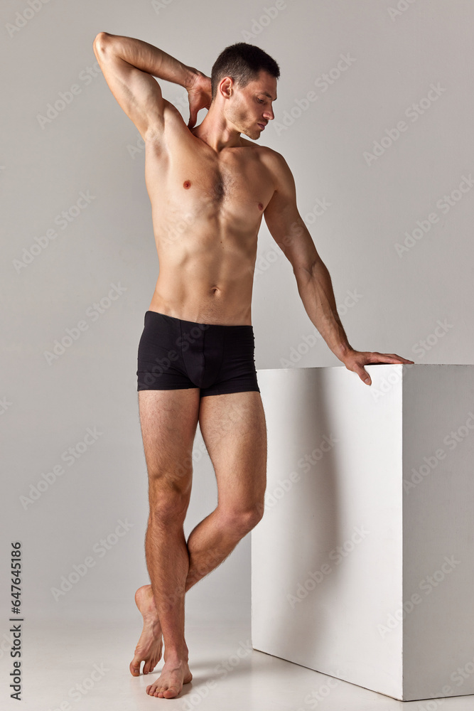 Full-length image of handsome, shirtless man with muscular body posing in underwear against grey studio background. Concept of men's health and beauty, body care, fitness, wellness, ad - obrazy, fototapety, plakaty 