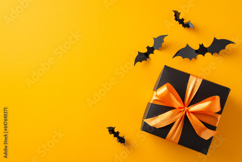 Share Halloween happiness with thoughtful gifts. Above shot captures woman's hands holding a black present box with a bow and paper bats on orange isolated background, with copyspace for ads or text