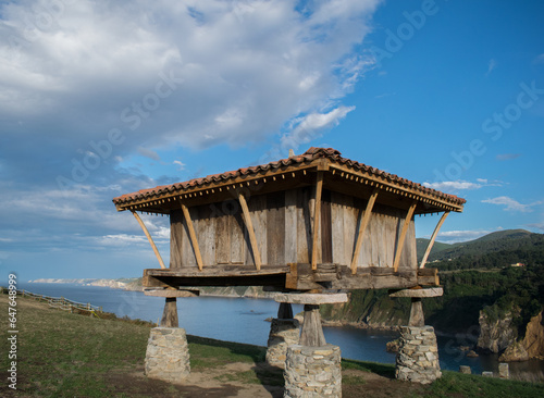 Hórreo asturiano a los pies de un acantilado con cielo azul y nubes