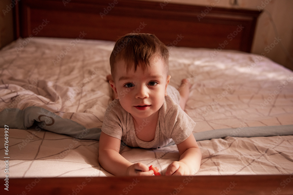One year old baby lies on his stomach on the bed in the morning in the sun. The child is funny, smiling, playing in a great mood. Care, furniture for infants. Close-up portrait of a boy. Lifestyle One year old baby lies on his stomach on the bed in the morning in the sun. The child is funny, smiling, playing in a great mood. Care, furniture for infants. Close-up portrait of a boy. Lifestyle