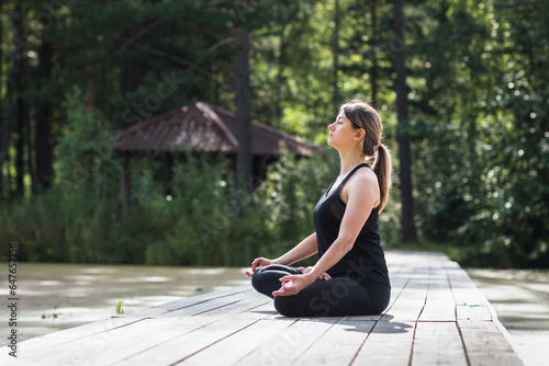 A woman is practicing meditation, sitting in a lotus position on a wooden bridge in a park on a summer sunny morning, doing yoga in black sportswear