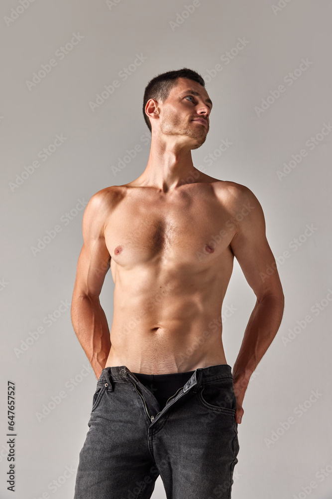 Portrait of young man with muscular body posing shirtless in jeans against grey studio background. Concept of men's health and beauty, body care, fitness, fashion, wellness, ad