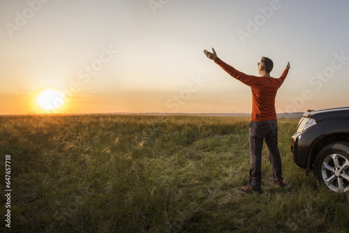 happy man tourist with open arms near his car at summer fieid at dawn or sunrise. Summer vacation adventure freedom auto travel concept. photo