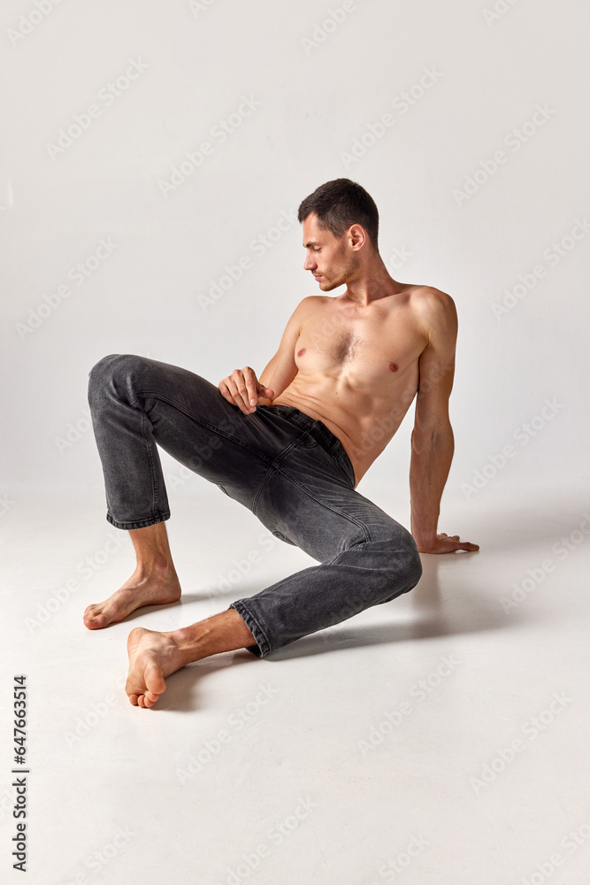 Portrait of handsome man in his 20s with muscular body shape posing shirtless in jeans against grey studio background. Concept of men's health and beauty, body care, fitness, wellness, ad
