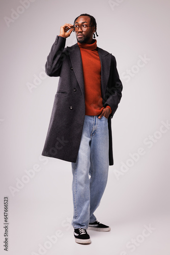 Fashionable young african american model posing in eyeglasses, classic coat and jeans on grey background