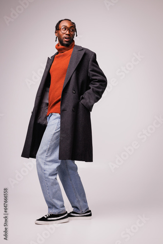 Confident african american man posing in eyeglasses, autumn coat and sneakers on grey background