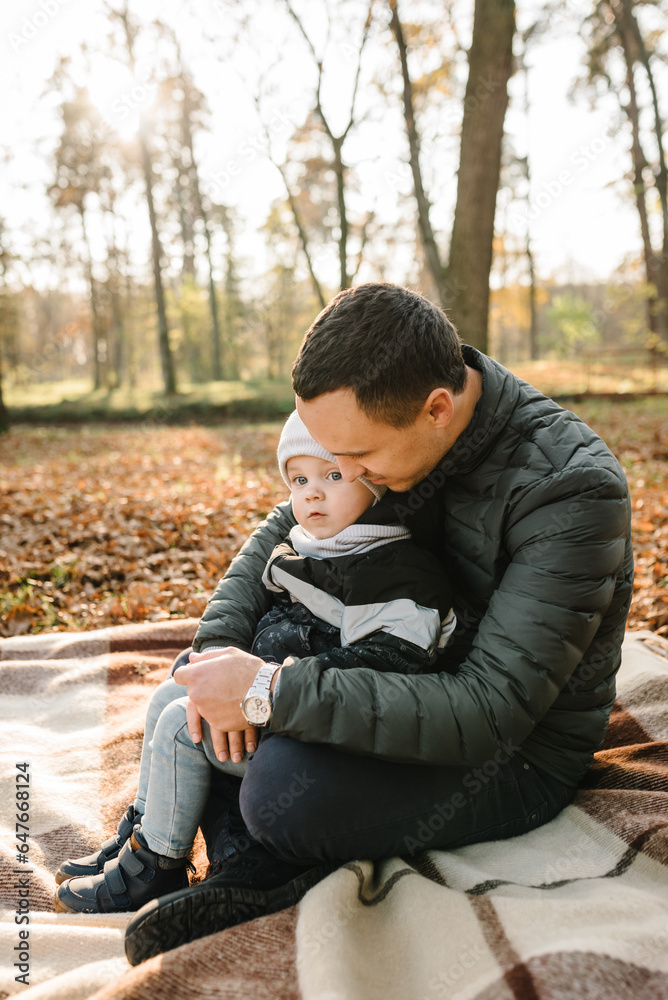 Dad embraces son child sitting on blanket on golden leaves in park. Father hugging kid have picnic in autumn forest in nature. Family holiday, spending time together at sunset. Autumn mood. Closeup. Dad embraces son child sitting on blanket on golden leaves in park. Father hugging kid have picnic in autumn forest in nature. Family holiday, spending time together at sunset. Autumn mood. Closeup.