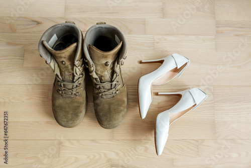 Old worn military boots, women's white shoes on wooden floor. Veteran and family concept.