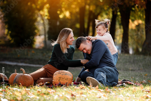 Thanksgiving day. Happy mom, dad and little child daughter having fun sit on grass with pumpkins at autumn park, enjoying spend time together at weekend. Parental care and happy carefree childhood