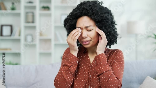 Tired african american female suffering from eye strain sitting on sofa at home. Sad black womant feels deterioration of vision, tension, closes her eyes and massages her eyeballs with her fingers photo