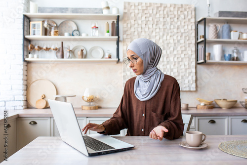 Young Muslim woman in hijab sitting worried in kitchen and using laptop, having technical problems, raising hands in frustration.
