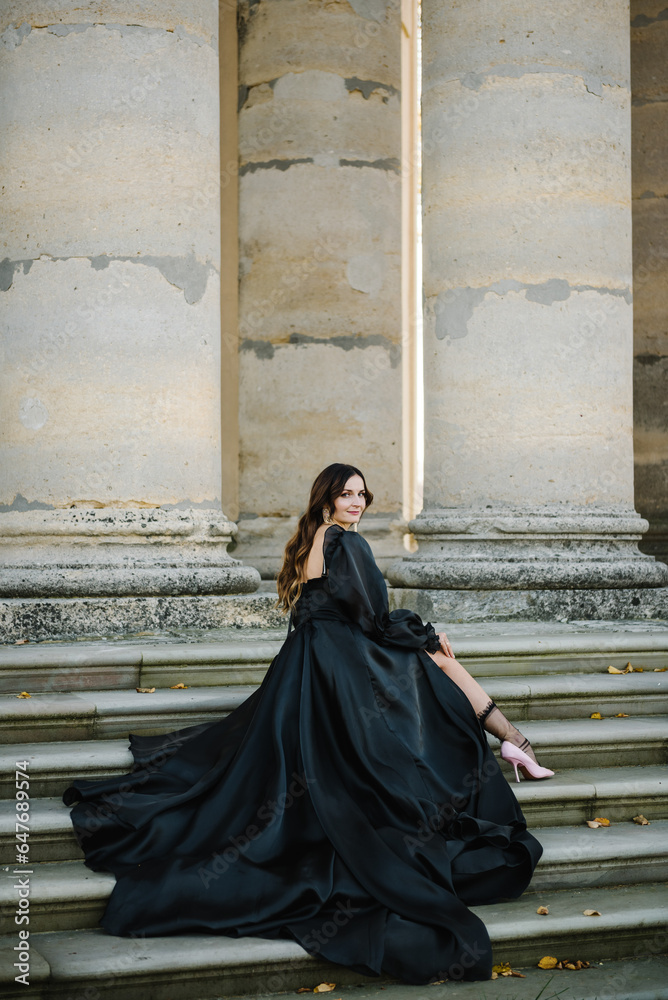 Naklejka premium Girl in black dress sitting on stairs near large columns of ancient temple at sunset. Female walk near Baroque Roman Catholic church in Pidhirtsi, Lviv, Ukraine. Woman in autumn day. Luxury bride