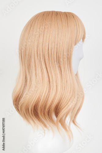 Natural looking blonde fair wig on white mannequin head. Middle length hair cut on the plastic wig holder isolated on white background, side view.