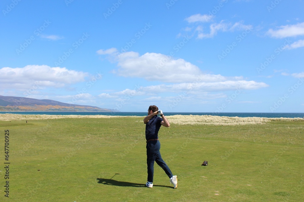 A young man swinging a golf club on a tee box surrounded by the beautiful views of the sea in the background, in Brora, Scotland A young man swinging a golf club on a tee box surrounded by the beautiful views of the sea in the background, in Brora, Scotland