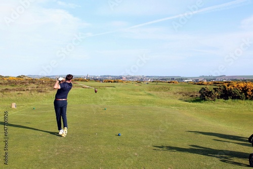 A young man swinging a golf club on a tee box surrounded by the beautiful views with St. Andrews in the background, in Scotland