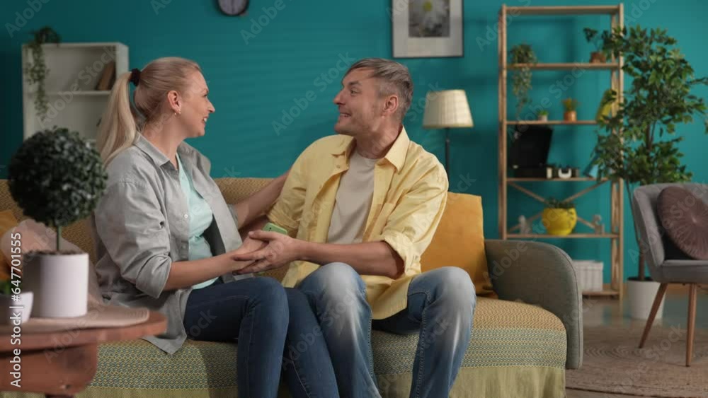 A woman is sitting on a sofa in the living room with a smartphone in her hands, a man comes up to her and sits next to her. The couple kisses, greeting, the man shares the good news, .