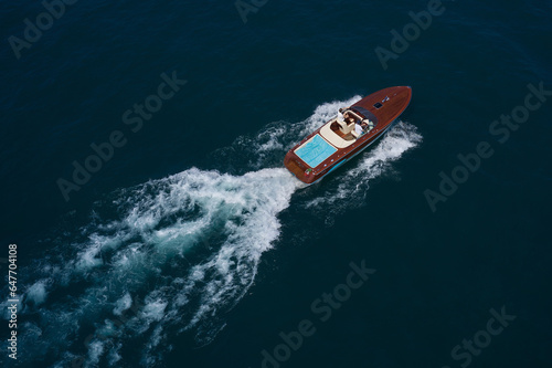 Luxury wooden big speedboat fast moving on dark water top view. Expensive wooden big boat with people moving on the water aerial view. Boat movement on the water.