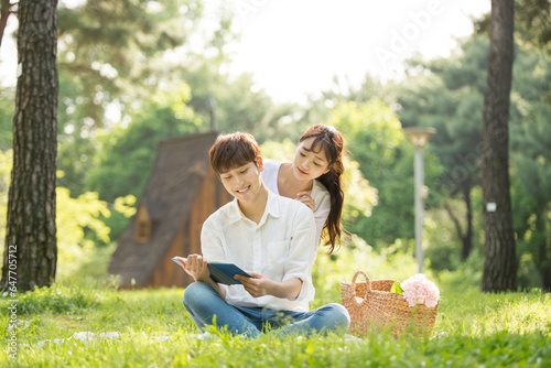 A young male and female couple are enjoying a pleasant picnic while drinking drinks and reading books on the forested park lawn. © PHOTOGRAPHER JH