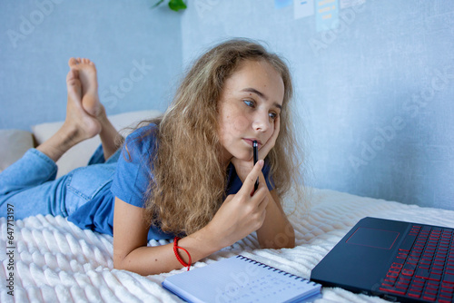 Blonde teenage girl on the bed doing homework using a laptop. Makes notes in a notebook. The concept of home education and self-study