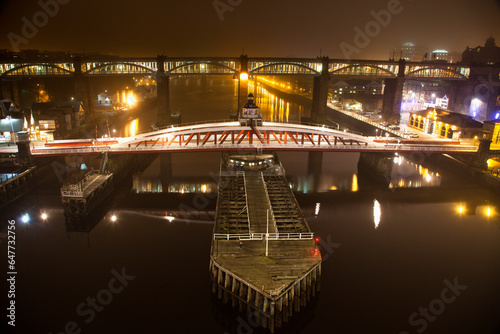 A Barge Passing Underneath A Bridge Travelling On The River Tyne Illuminated At Night; Newcastle, Northumberland, England